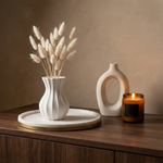 Decorative setup with a white vase, candle, and dried plants on a wooden surface.
