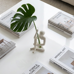 Overhead view of a white seated figure vase holding a large split green leaf, surrounded by books on a white table.