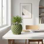 Green textured vase with a plant on a white table in a bright room.