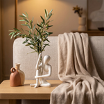 White seated figure vase holding leafy branches beside a small two-tone vase and a sofa draped with a blanket.