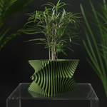 Green ribbed square planter with a leafy plant on a clear stand, framed by foliage against a dark background.
