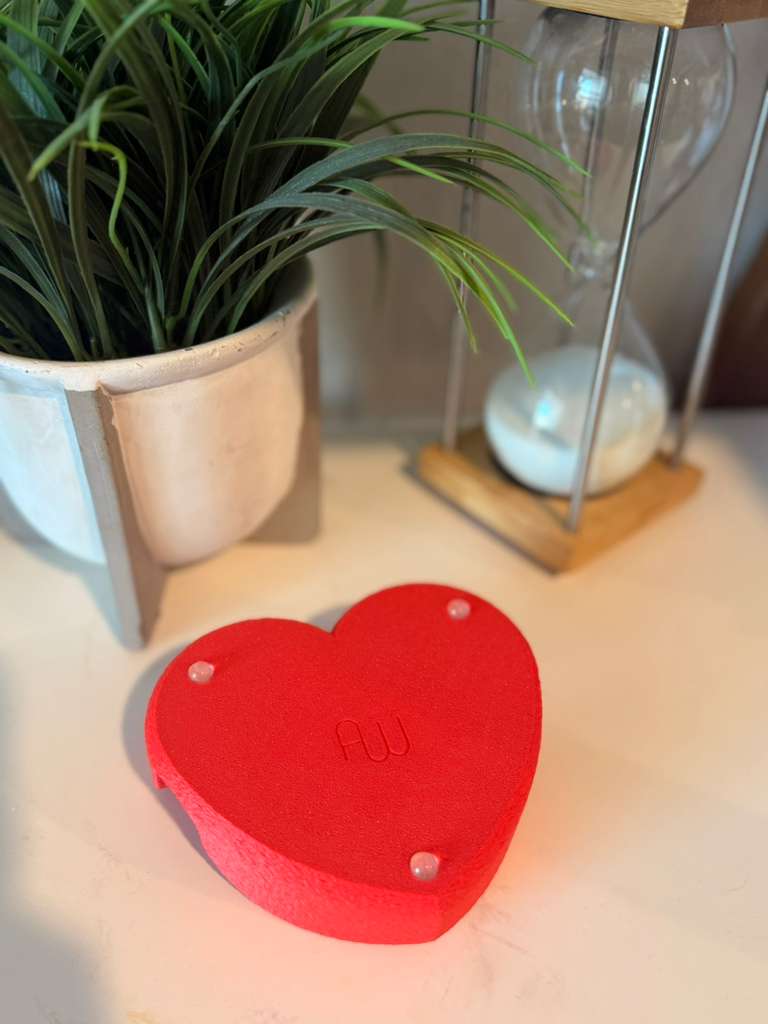 Red heart-shaped object with 'AU' branding on a light surface, next to a potted plant and hourglass.