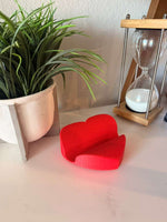 Red heart-shaped object on a surface with a plant and hourglass in the background
