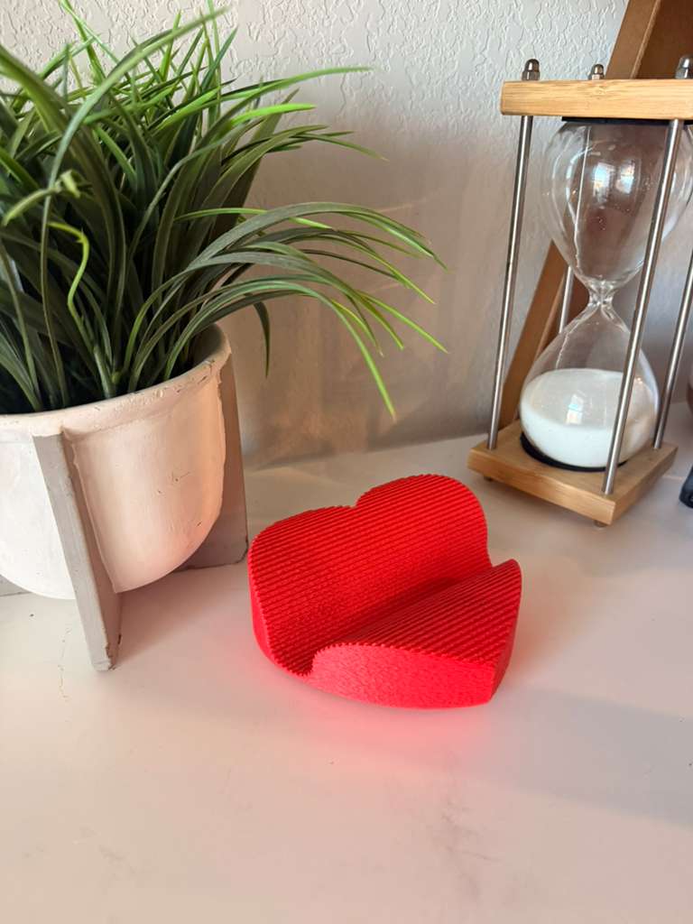 Red heart-shaped object on a surface with a plant and hourglass in the background