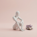 White textured seated NObody Reflection figure beside a purple crystal cluster on a blush pink background
