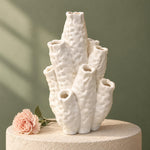 White coral-shaped vase with bumpy tubular branches on a round pedestal beside a pink flower, against a green background