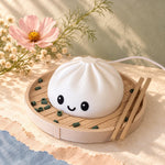 Cute dumpling lamp, a white kawaii decor night light with a smiling face, sits in a steamer basket with chopsticks beside pink flowers on a cream surface.