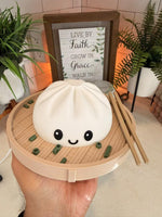 Hand holding the smiling white dumpling lamp in its steamer basket with chopsticks, a small table lamp showing its palm-sized scale, in front of a framed sign and green plants.