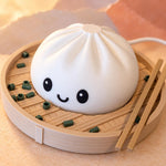 Close-up of the smiling white dumpling lamp, a cute small table lamp with pleated top, in a steamer basket with chopsticks and a power cord on a peach backdrop.