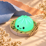 Smiling dumpling lamp glowing bright green, a cute color-changing kids night light in a steamer basket with chopsticks on peach paper with blue cloth and dried flowers.