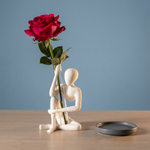 White seated figure vase holding a clear tube with a red rose, beside a small gray dish on a wooden table.