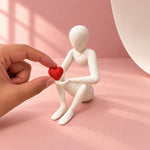 Hand holding the red heart beside the seated white Love’s Embrace figure on pink paper