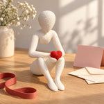 White Love’s Embrace figure holding a red heart beside a note and ribbon on a wooden table