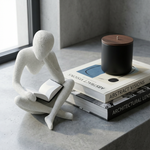 White sculpture of a person reading a book on top of books with a black candle on a concrete surface.