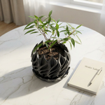 Black wavy pot with a plant on a marble surface next to a notebook and pen.