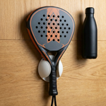 Black and orange padel racket in a white wall mount on wood paneling beside a black bottle.