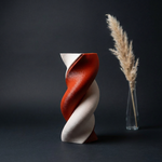 Rust-red and cream twisting vase beside a narrow clear vase with a dried grass stem against a dark background.