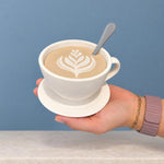 White coffee cup storage container with latte-art lid and spoon held against a blue wall