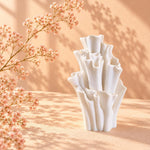 White matte textured sculptural vase with ruffled coral-like tubes on a sunlit peach surface, with dried blossom sprigs and soft leaf shadows on the wall.
