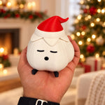 Hand holding a small garlic lamp with a matte textured ivory body, red elf hat and black feet in front of a blurred, glowing christmas tree. A garlic lamp christmas light shown unlit.
