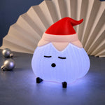 Ridged garlic lamp glowing soft white-blue, wearing a red elf hat with a pale pink cuff, in front of a fanned paper backdrop on a dark surface. A glowing christmas light.