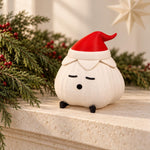 Elf hat garlic lamp with a matte ridged ivory body and red hat on a textured cream mantel beside pine branches with red berries and a pale paper star. A christmas light shown unlit.