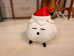 Everyday photo of a garlic lamp with a matte ivory textured body, red elf hat and black feet on a light countertop, with a white USB cord, a wooden chair and a small framed sign behind it. Shown unlit.