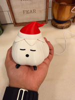Hand holding a garlic lamp with a textured ivory body, red elf hat and black feet over a kitchen counter, with a framed sign and a wooden container behind it. A christmas light shown unlit.