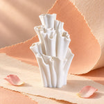 White matte textured coral-shaped bud vase with ruffled tubular openings, a sculptural vase about 7 inches (18 cm) tall, on peach paper with scattered pink petals.
