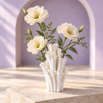Cream flowers with green leaves in a white matte textured bud vase with ruffled tubes, on a pale tan block in front of a lavender arched wall.