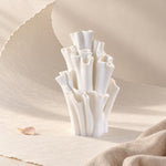 White matte textured sculptural vase with ruffled, wavy tubes on a cream surface with beige linen fabric and a few pink petals, in soft side light.
