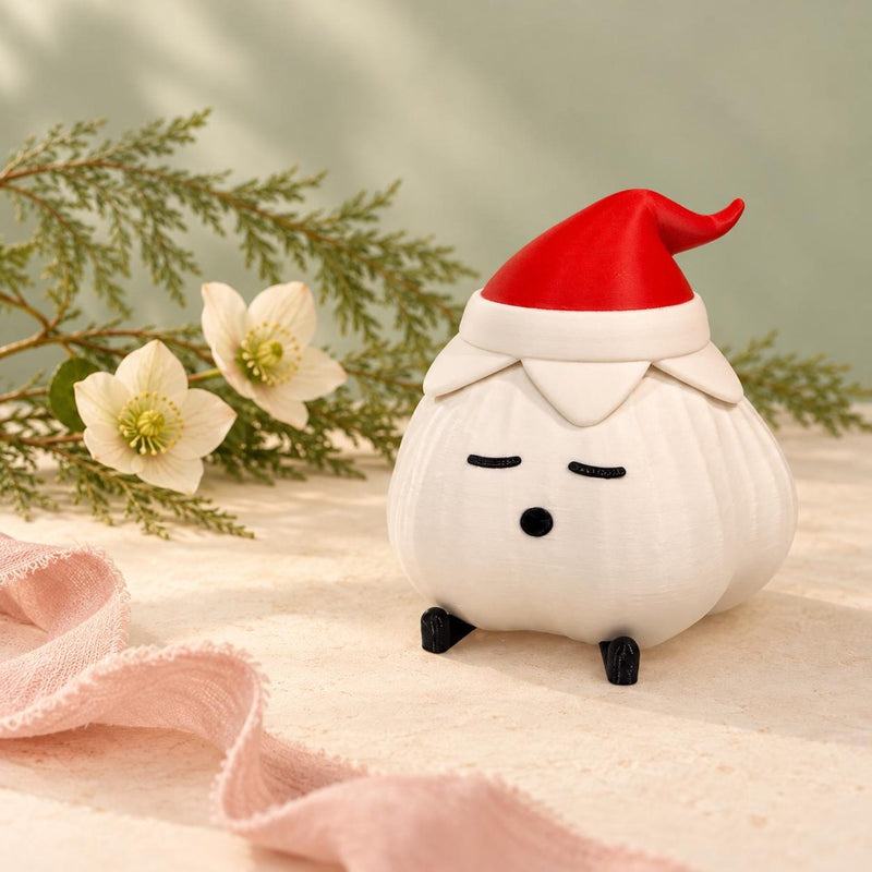 Elf hat garlic lamp with a textured ivory body, red pointed hat and black feet on cream linen with pale hellebore flowers, pine sprigs and pink fabric against a sage-green wall. A soft christmas light for a winter shelf.