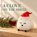 Garlic lamp in a red elf hat with a matte textured ivory body on a light wood shelf beside pine and a red bow, with bold headline text above it. A festive garlic lamp for holiday shelves.