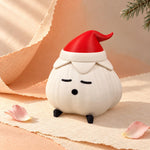 Garlic lamp with a matte textured ivory body, red elf hat and black feet on a cream doily with a pink rose petal and pine sprig against a peach backdrop. A cozy christmas light shown unlit.