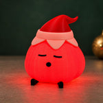 Garlic lamp glowing bright red, wearing a red elf hat with a pale pink cuff, on a dark surface with a gold ornament and dark green background. A festive garlic lamp christmas light.