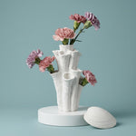 White coral-shaped vase with pink and purple flowers on a round white stand beside a seashell