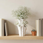 White coral-shaped vase filled with tiny white flowers on a wooden shelf between books and a small potted plant