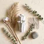 Flat lay of a white coral-shaped vase surrounded by dried grasses, a cotton stem, green sprigs, fabric and a stone