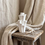 Empty white coral-shaped vase on a weathered wooden stool beside shells and draped beige fabric
