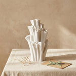 Empty white coral-shaped vase on a fabric-covered table beside dried flowers and an envelope tied with green ribbon