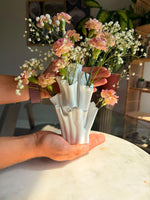 Hands holding a white coral-shaped vase filled with pink and peach flowers and tiny white blossoms