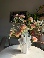 White coral-shaped vase with pink and peach flowers and tiny white blossoms on a round table in a sunlit room
