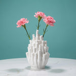 White coral-shaped vase with clustered narrow tubes and pink flowers against a teal background
